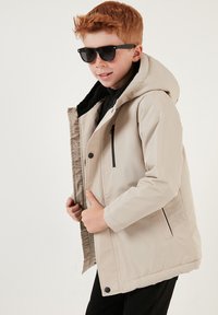 Beige isolerad jacka med huva, dragkedjestängning och svarta accenter. Har en mjuk, texturerad innervaddering och sidofickor.