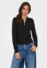 Czarna dzianinowa kardigan z zamkiem błyskawicznym, z wzorem w warkocze, z małym akcentem czerwonego serduszka na piersi. Połączona z niebieskimi dżinsami.