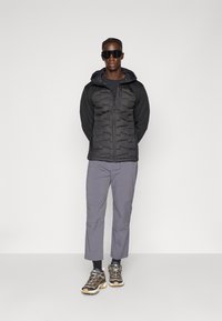 Black padded jacket with a dark hood, gray pants, and chunky black and tan sneakers. Sunglasses complete the outfit.