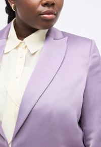 Γυναίκα που φοράει χρυσούς κρίκους ως σκουλαρίκια, κρεμ πουκάμισο με κουμπιά και μωβ σακάκι, γυρισμένη ελαφρώς προς τα αριστερά μπρο στά σε απλό λευκό φόντο.