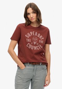 Camiseta de algodón color marrón con escote redondo y mangas cortas. Presenta un estampado gráfico en gris claro, el texto dice "SUPERDRY TALL PINE COUNCIL."