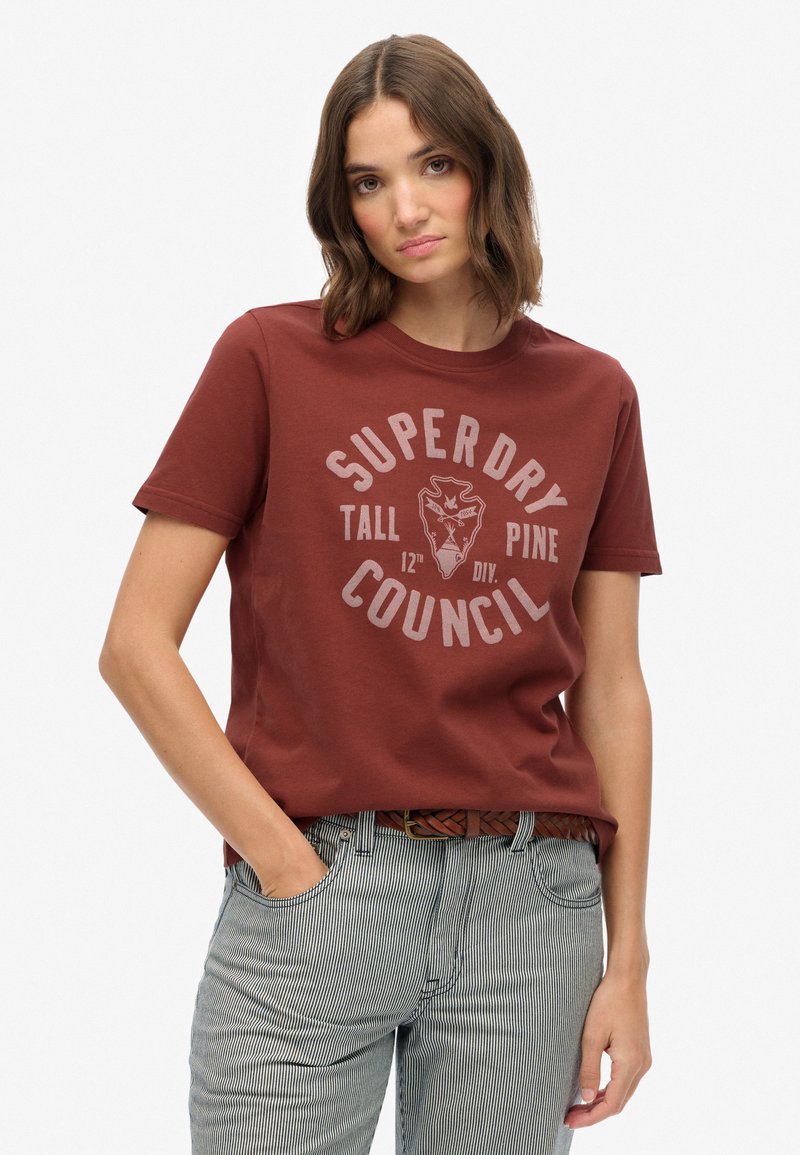 Camiseta de algodón color marrón con escote redondo y mangas cortas. Presenta un estampado gráfico en gris claro, el texto dice "SUPERDRY TALL PINE COUNCIL."