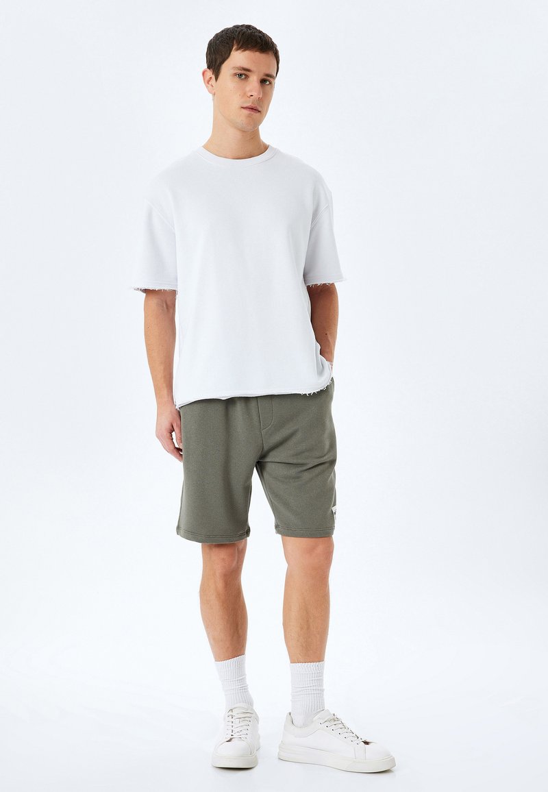 Ung mand står med den ene hånd i lommen, iført en hvid oversized skjorte med kortærmer, olivengrønne shorts, hvide sokker og hvide sneakers.