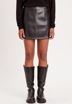Mini-jupe en simili cuir noir avec une texture lisse, fentes sur les côtés et couture contrastante. Associée à des bottes noires ajustées jusqu'aux genoux.