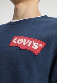 Námořnická modrá mikina s kulatým výstřihem, s výrazným červeným vyšitým logem Levi's na hrudi, klasický střih a strukturovaná tkanina.