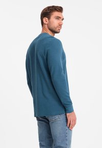 Mann, der sich leicht dreht, trägt ein langärmliges türkisfarbenes Hemd und hellblaue Jeans und steht vor einem schlichten weißen Hintergrund.