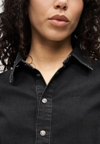 Μαύρο πουκάμισο από τζιν με κουμπιά, αντίθετη ραφή και ασημένιες μεταλλικές λεπτομέρειες. Διαθέτει κλασικό γιακά και διακριτική λεπτομέρεια με λογότυπο.