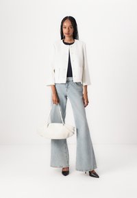 Weiß strukturierte Jacke mit Dreiviertelärmeln, kombiniert mit hellblauen Schlagjeans. Das Model hält eine weiße Handtasche und trägt schwarze Schuhe.