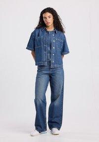 Giacca di jeans di un blu medio con maniche corte, dotata di due tasche frontali e bottoni argentati. Abbinata a jeans a gamba larga, anch'essi blu.