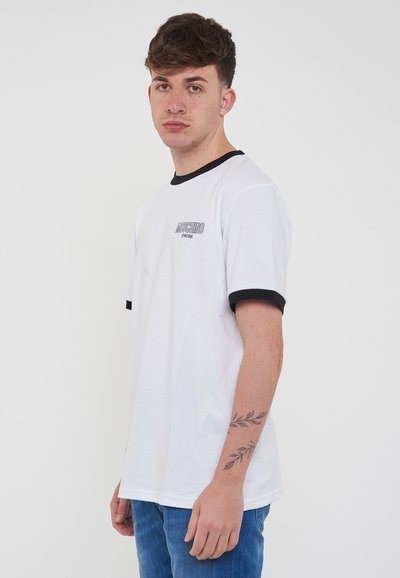 T-shirt bianca a maniche corte con colletto e polsini a coste neri, con un logo stampato sul fronte. Il tessuto sembra liscio e morbido.