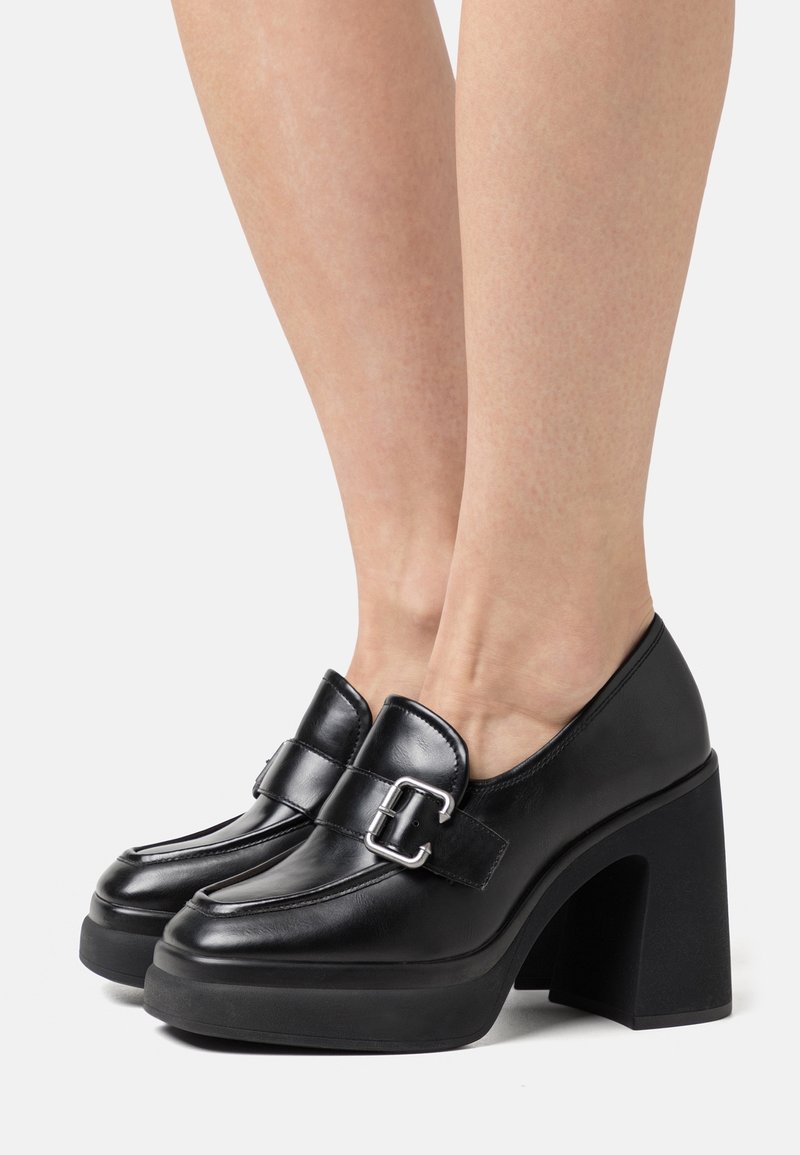 Escarpins à plateforme en cuir noir avec détail de boucle argentée portés aux pieds sur un fond uni.