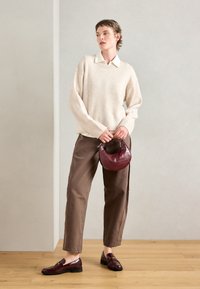 Beige strikkegenser, brune litt løse bukser, mørkerød teksturert håndveske og blanke mørkerøde loafers på et tregulv. Nøytral bakgrunn.