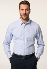 Camisa de manga larga azul claro con cuello abotonado, de tela texturizada, combinada con pantalones oscuros y un cinturón negro. El modelo está de pie con las manos en las caderas.