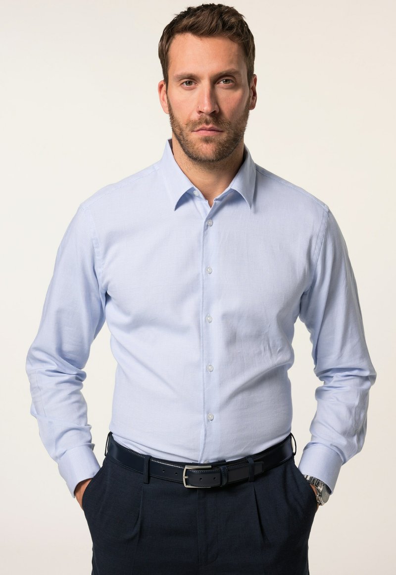 Camisa de manga larga azul claro con cuello abotonado, de tela texturizada, combinada con pantalones oscuros y un cinturón negro. El modelo está de pie con las manos en las caderas.