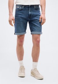 Άνδρας που φοράει σκούρες μπλε τζιν βερμούδες με ρεβέρ, λευκές κάλτσες, μπεζ αθλητικά παπούτσια και λευκό πουκάμισο, στέκεται μπροστά σε ένα απλό φωτεινό φόντο.