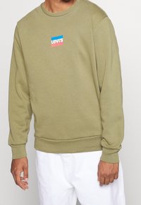 Sweat-shirt vert olive, col rond, tissu doux, poignets et ourlet côtelés, avec un logo rectangulaire Levi's en bleu et rose.