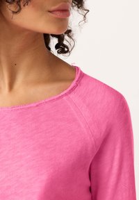 Pribličen posnetek ženske vratu in spodnjega dela obraza s kodrastimi lasmi, ki nosi rožnato zgornjo majico z okroglim izrezom in rahlo raztrganimi robovi.