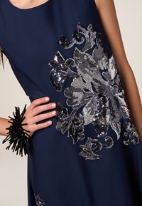 Marineblaues Kleid mit großen silbernen und schwarzen floralen Paillettenverzierungen. Das Modell trägt ein schwarzes, strukturiertes Armband, das das Design akzentuiert.