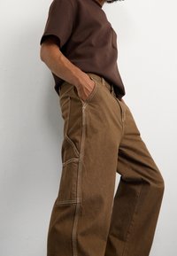 Pantalon marron à jambes larges en tissu texturé, avec surpiqûres contrastantes, poches latérales et coupe décontractée, associé à une chemise marron.