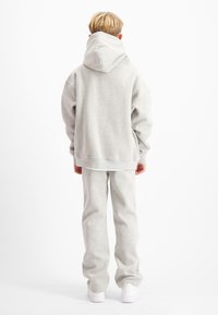 Persoon met kort blond haar, staand, in een lichtgrijze hoodie en bijpassende joggingbroek, van achteren gezien op een witte achtergrond.