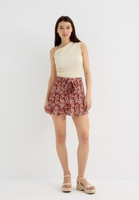 Top de textura cremosa con un tirante en un hombro, combinado con una falda roja con flores que tiene un dobladillo con volantes y un lazo en la cintura. Sandalias de cuña.