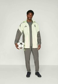 Veste de sport vert clair et grise avec des accents noirs, pantalon gris assorti et chaussures de sport noires. Tenant un ballon de soccer noir et blanc.