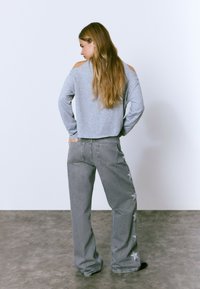 Woman with long hair wearing a gray cold-shoulder top and wide-leg gray jeans with star designs, standing with hands in back pockets.