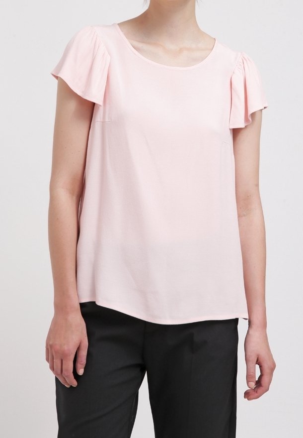 Blouse rose clair à manches courtes avec un col rond et des manches volantes, fabriquée en tissu lisse, portée avec un pantalon sombre.