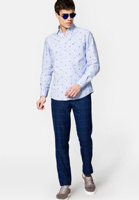 Jasnoniebieska koszula z guzikami z małym nadrukiem łodzi żaglowej, w połączeniu z ciemnymi spodniami w kratę i szarymi sneakersami. Okulary przeciwsłoneczne dopełniają stylizację.