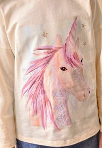 Chemise à manches longues couleur crème avec un motif de licorne arborant une crinière rose, des accents d'étoiles en paillettes et des sequins sur le visage du cheval.