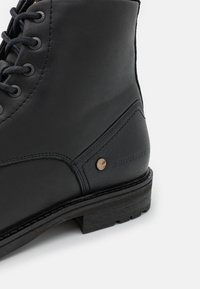 Černá kožená kotníková bota s kulatými tkaničkami, kovovými kroužky, texturovanými detaily a jemným logem poblíž paty na dřevěné podrážce.