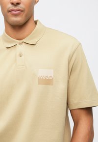 Polo shirt beige en tissu texturé, avec un petit logo carré en beige plus clair. Patte de boutonnage à trois boutons et manches courtes.