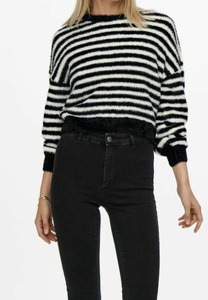 Vrouw draagt een zwart-witte gestreepte, pluizige trui, ingestopt in een zwart, hooggetailleerd jeans, staand met de armen ontspannen langs haar lichaam.