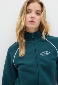 Chaqueta con cremallera de color teal oscuro con ribetes blancos. Presenta un cuello alto y texto bordado "PARIS 1996 Rue de Rivoli SPORT CLUB". Textura suave.
