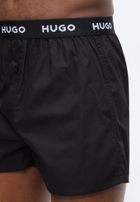 Μαύρα κοντά μπόξερ με κουμπωτό άνοιγμα και ελαστική μέση με λευκά λογότυπα "HUGO" στη μέση και το πάνω μέρος των μηρών.