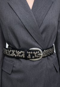 Črni usnjeni pas okrašen s srebrnimi kovinskimi zakovicami, ki ima veliko srebrno sponko; nošen čez sivi blazer z teksturirano površino.