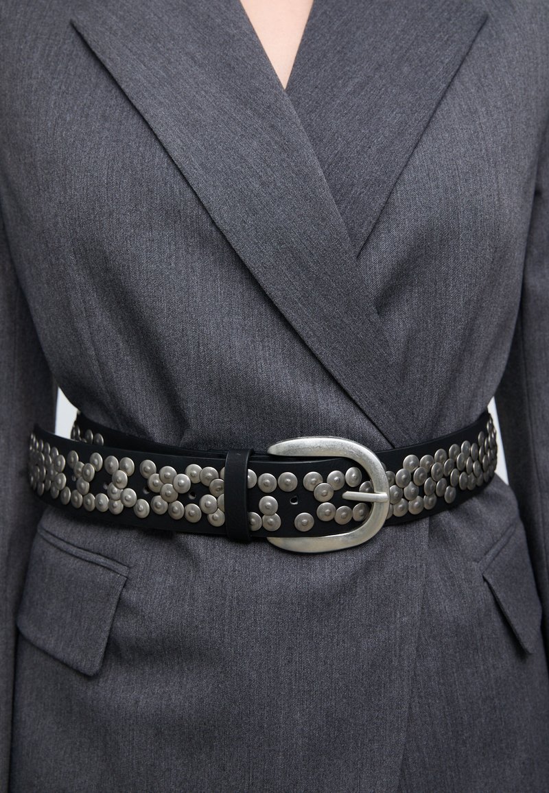 Črni usnjeni pas okrašen s srebrnimi kovinskimi zakovicami, ki ima veliko srebrno sponko; nošen čez sivi blazer z teksturirano površino.