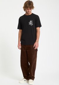 Man wearing black T-shirt with a small graphic on chest, brown loose pants, and white sneakers, standing and looking down.