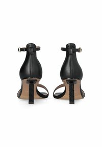 Sandalias de tacón de cuero negro con correa en el tobillo, parte trasera abierta y tacones decorativos con un patrón geométrico.