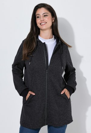 Vrouw met lang bruin haar, dragend een donker gestructureerde hoodie met rits over een wit shirt, handen in de zakken, glimlachend en opzij kijkend.