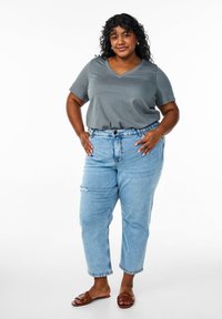T-shirt grigio a scollo a V, jeans azzurri leggermente strappati e sandali marroni. Capelli ricci, mani sui fianchi, sfondo semplice.