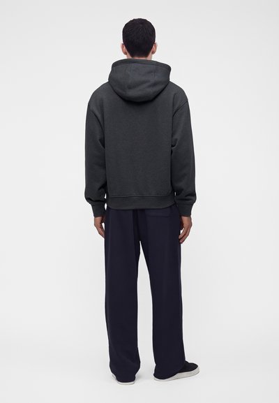 Homme vu de dos portant un sweat à capuche gris foncé, un pantalon large bleu marine et des baskets noires sur un fond clair uni.