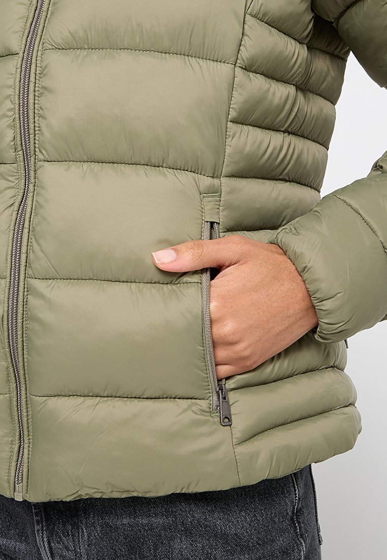 Olijfgroene pufferjack met een ritssluiting, voorzien van horizontale naden en een zijzak, gemaakt van lichtgewicht, gestructureerd materiaal.