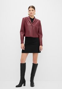 Giacca cropped finta pelle colore bordeaux con colletto e una tasca, abbinata a una gonna nera aderente e stivali neri con tacco al ginocchio.