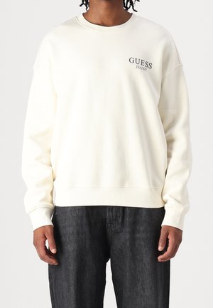 Cremegul sweatshirt med ribbede manchetter og kant, med "GUESS JEANS" logo broderet i mørkegrå på venstre bryst. Bæres med sorte jeans.