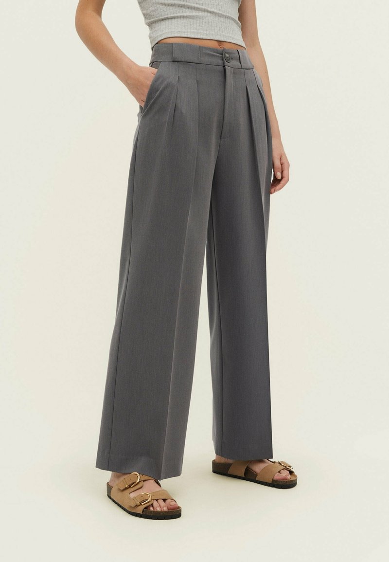 Pantalons plissés gris à taille haute avec poches latérales, présentant une fermeture à bouton et un design ample à jambes larges, associés à des sandales marron.