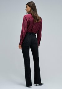 Camisa de satén burdeos con mangas largas y detalles de botones, combinada con pantalones vaqueros acampanados de tiro alto en negro y zapatos de tacón negros.