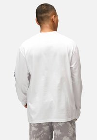 Maglietta bianca a maniche lunghe, in tessuto di cotone, colletto rotondo, orlo dritto. Pantaloni grigi tie-dye sotto, senza grafiche o dettagli visibili.