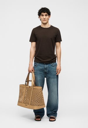 Noormees seisab, hoides suurt punutud beežikat kotti, kannab tumedat pruuni t-särki, lahtisi siniseid teksaseid ja musti sandaale tavalise tausta ees.