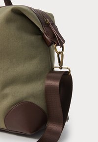 Lerretsbag i oliven grønn med brune skinnaksenter. Har en glidelås, strukturert overflate og en justerbar stropp med metallkrok.
